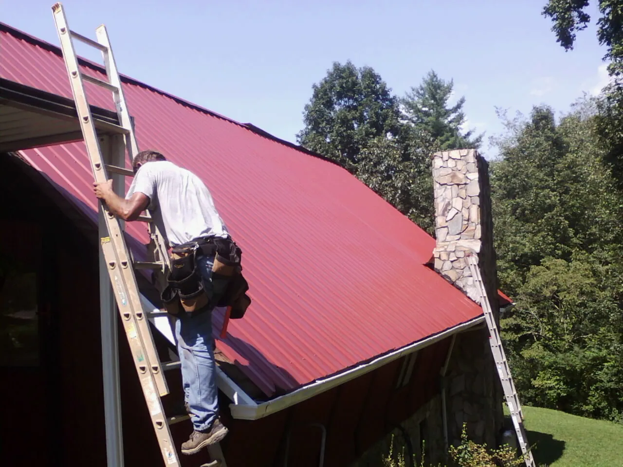 Roofer on a ladder beside a red metal roof and stone chimney — Soffit & Fascia Repair by Roofing Brothers in Laurel Hill